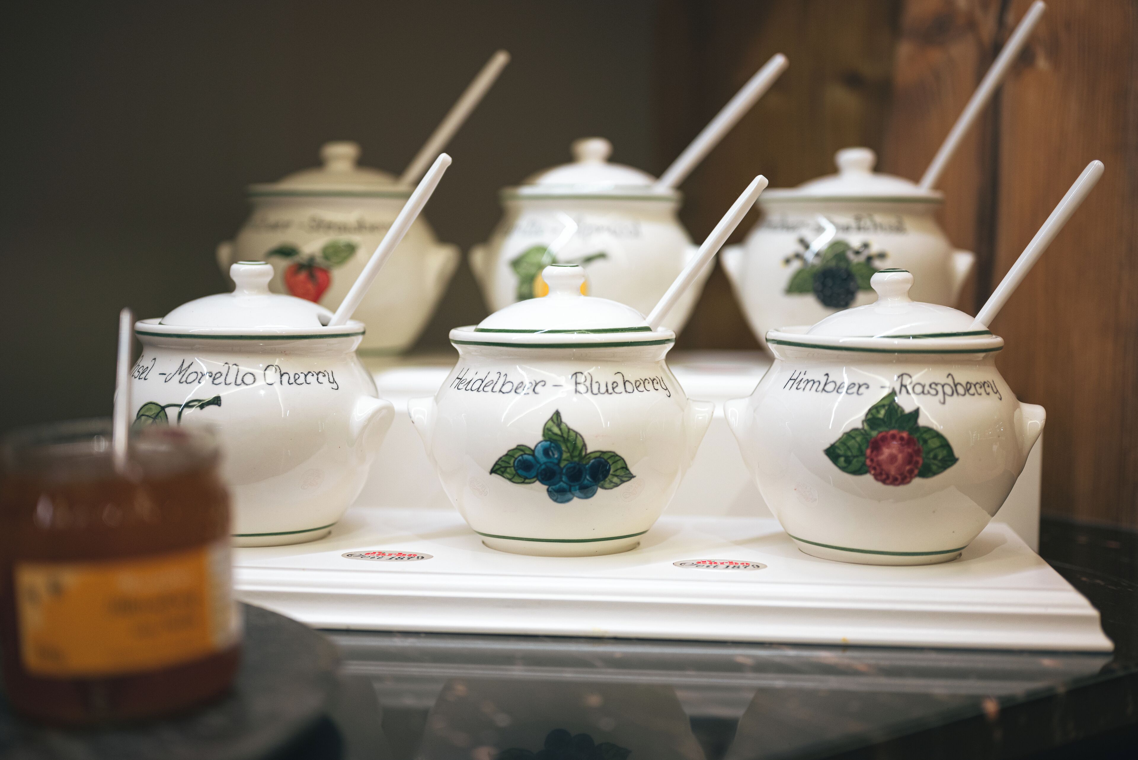 A set of white ceramic containers with green trim and labels for different types of jam. Each container has a white spoon inserted into it.