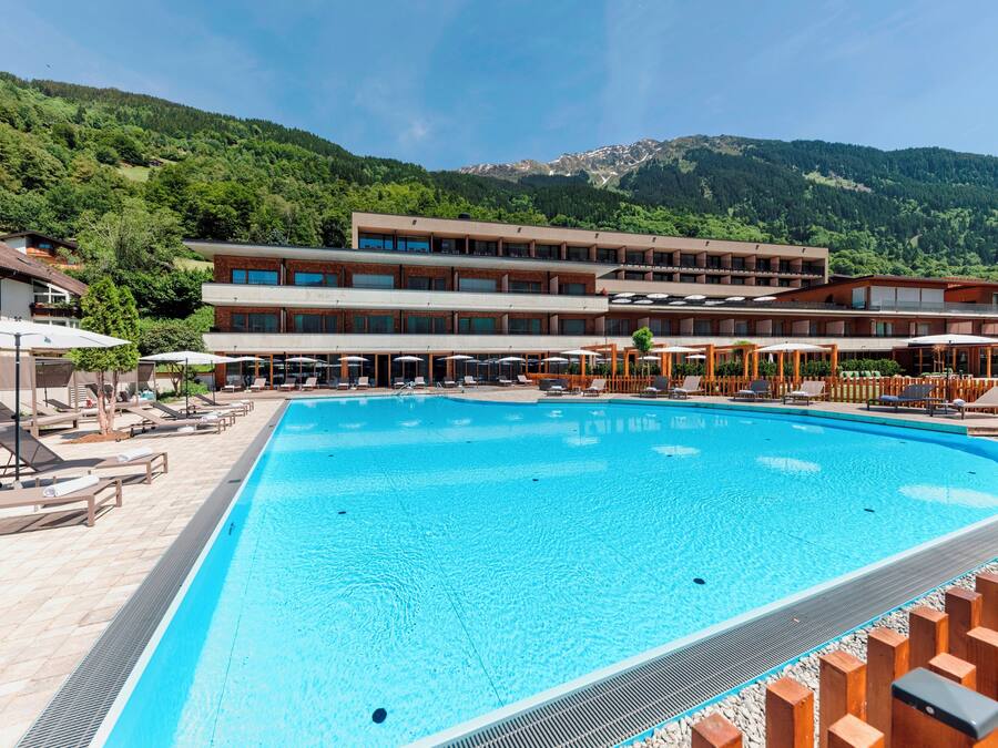 En swimmingpool med solstolar och parasoller, omgiven av ett hotell med flera våningar och en bergig kuliss i bakgrunden.