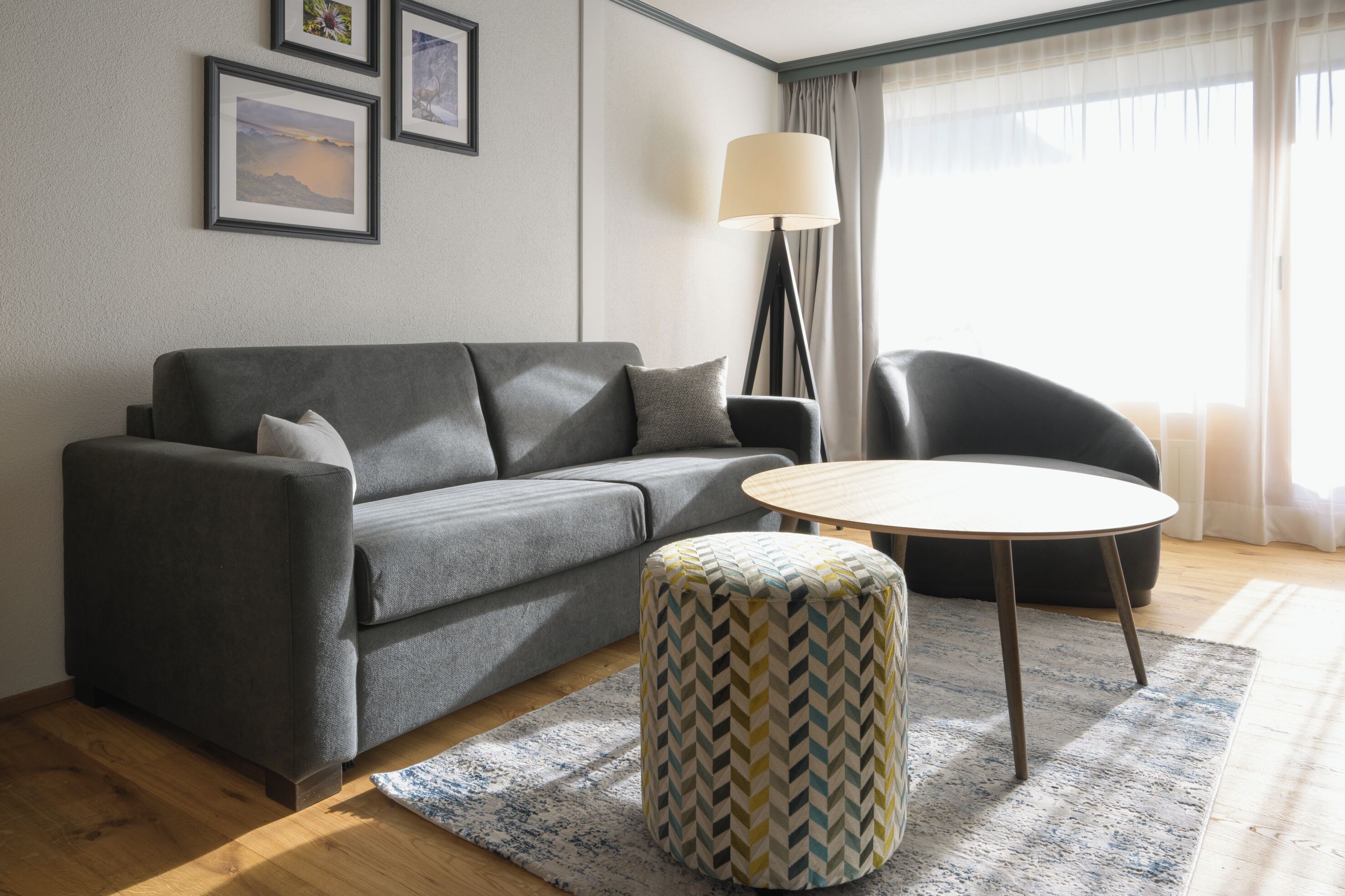 Una sala moderna con un sofá gris, una mesa de centro redonda, un reposapiés estampado, una lámpara de pie y una silla.