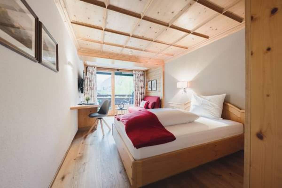 A bedroom with a wooden ceiling, a bed with white and red bedding, a desk with a chair, and a window with curtains.