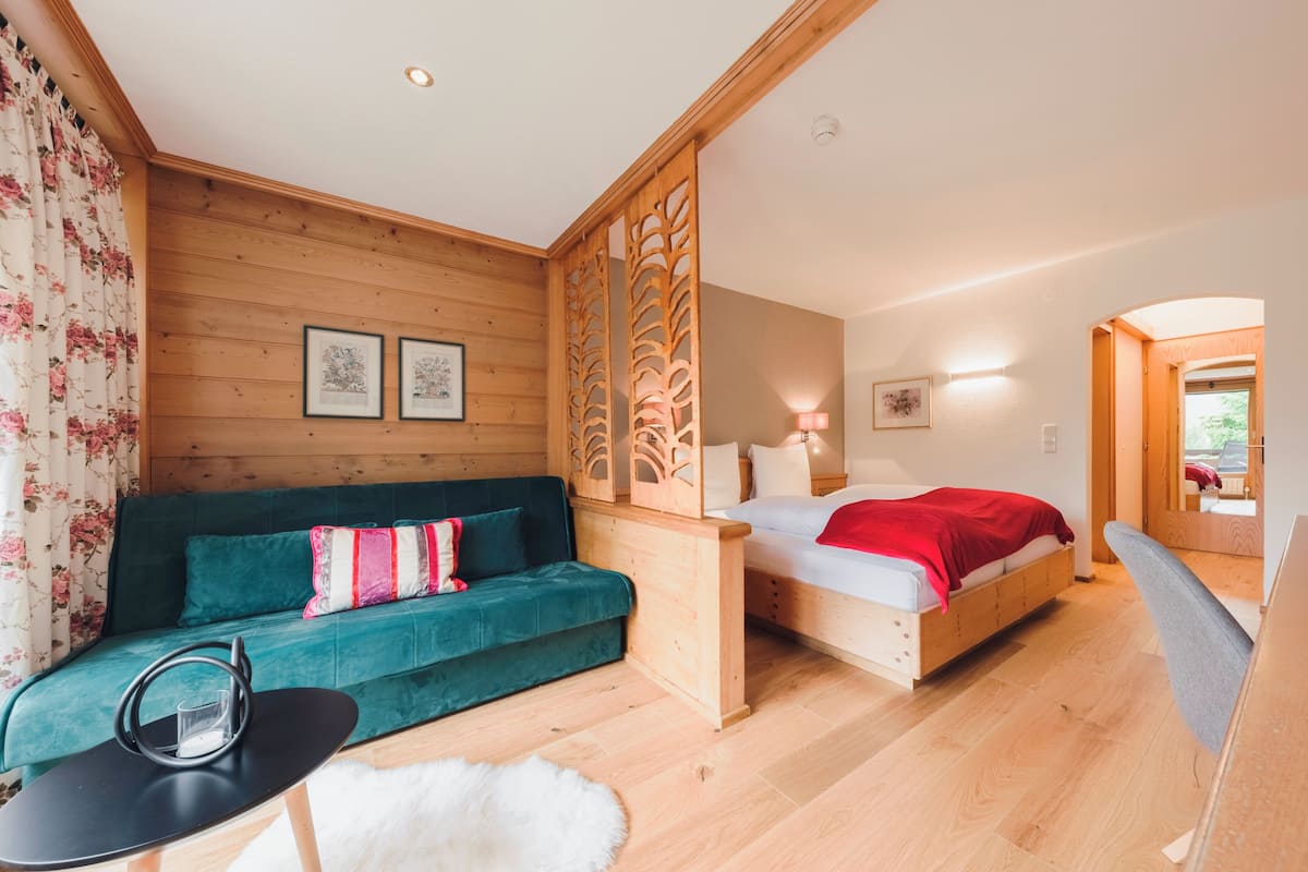 A room with a wooden wall, a green sofa, a bed with a red blanket, and a small table with a glass.