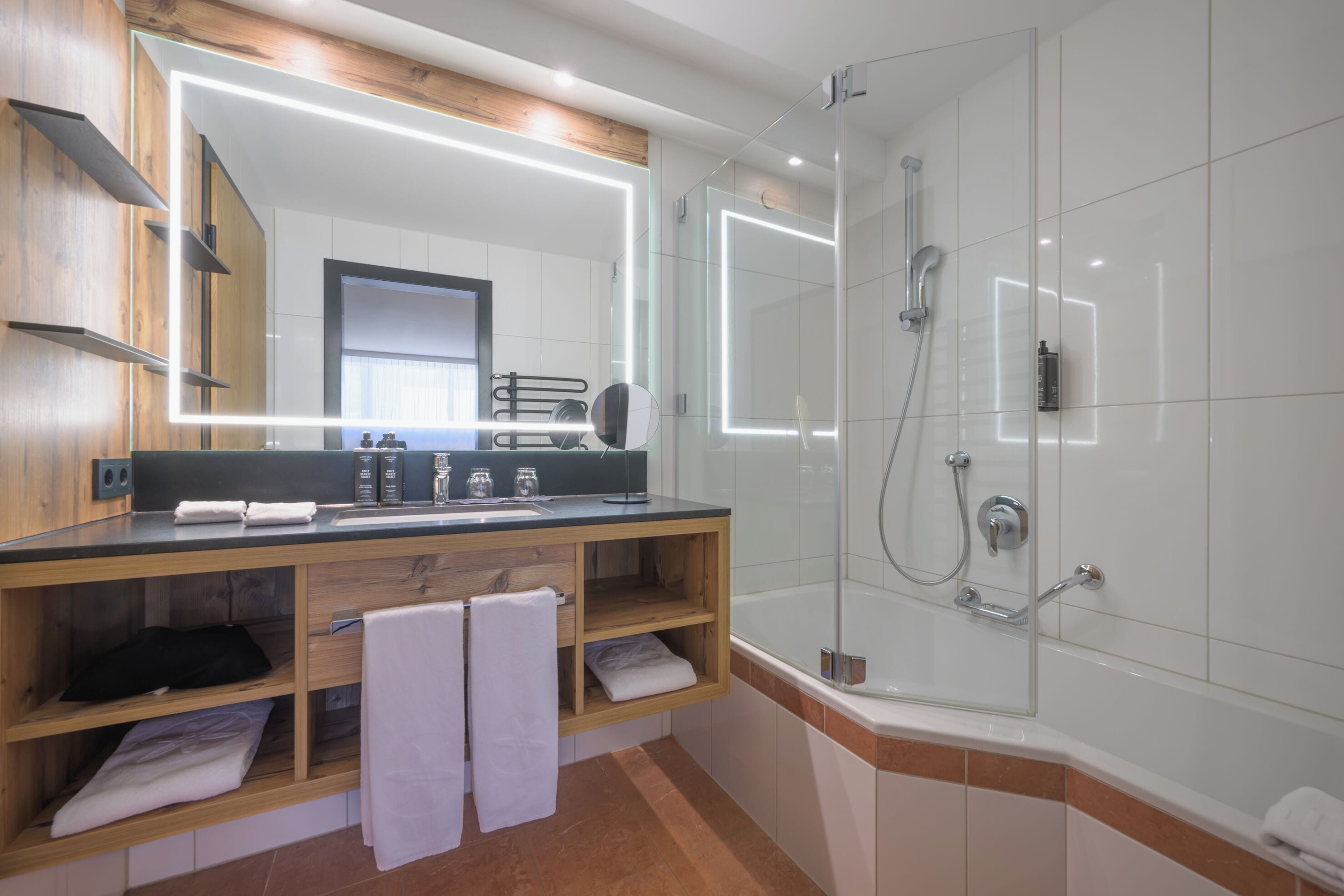 Un baño moderno con ducha de vidrio, tocador con espejo y repisa de madera con toallas.