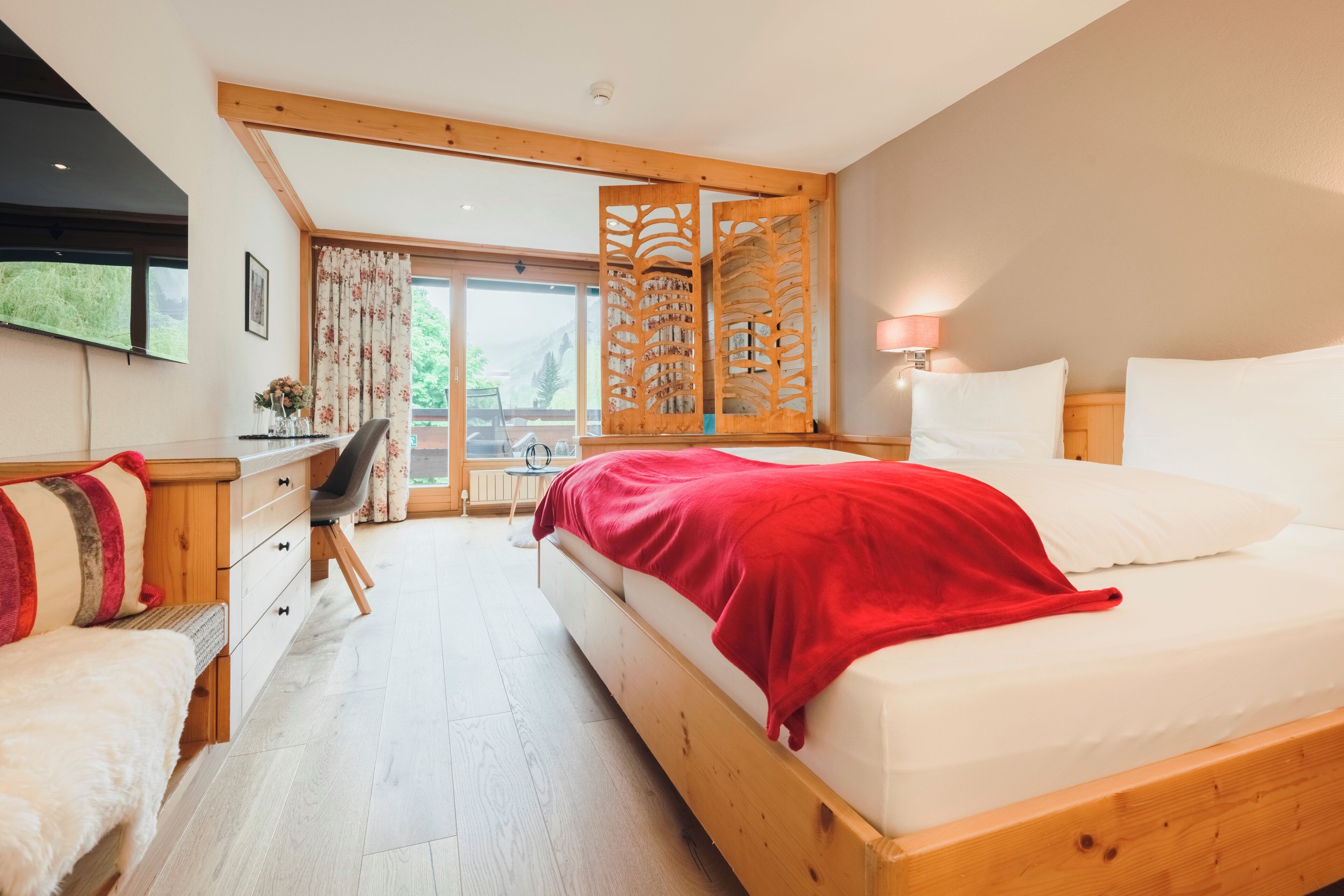 A modern bedroom with a large bed, wooden headboard, a desk with a chair, a TV mounted on the wall, and a view of the outdoors through a glass door.