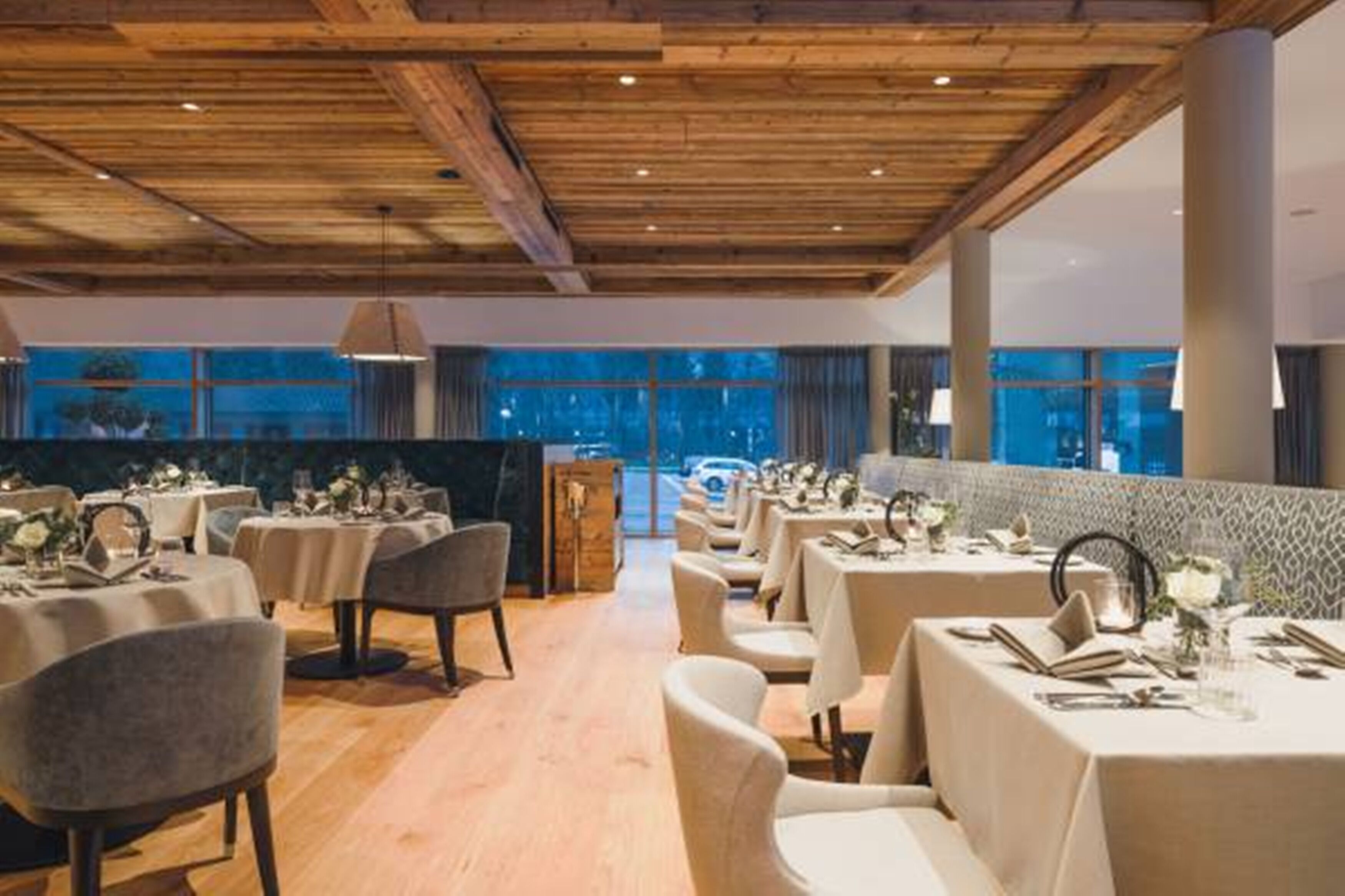 Ein Restaurant mit Holzbalkendecke, gedeckten Tischen mit weißen Tischdecken und großen Fenstern mit Blick nach außen.