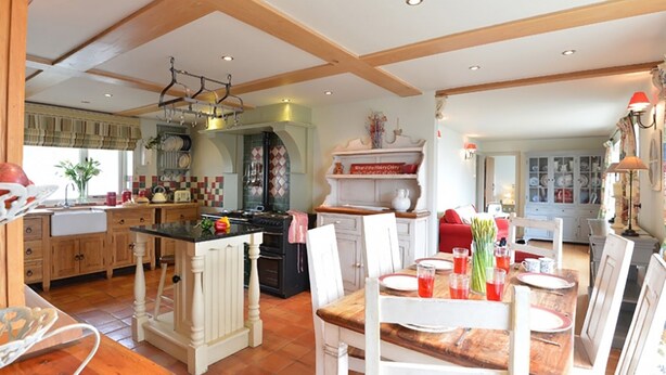 Une cuisine spacieuse avec des poutres apparentes, un îlot central et un coin repas avec une table dressée pour quatre personnes.
