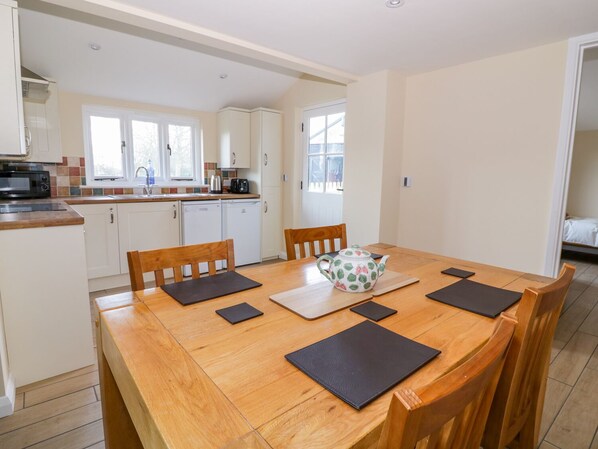 A modern kitchen with white cabinets, a microwave, and a sink. There is a dining table with four chairs and a teapot. - The Granary (Halstead)