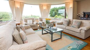 A spacious living room with a large sofa, two armchairs, a coffee table, and a ceiling fan. There are large windows offering a view of greenery outside. - Bryn Y Mor, Borth Y Gest (Porthmadog)
