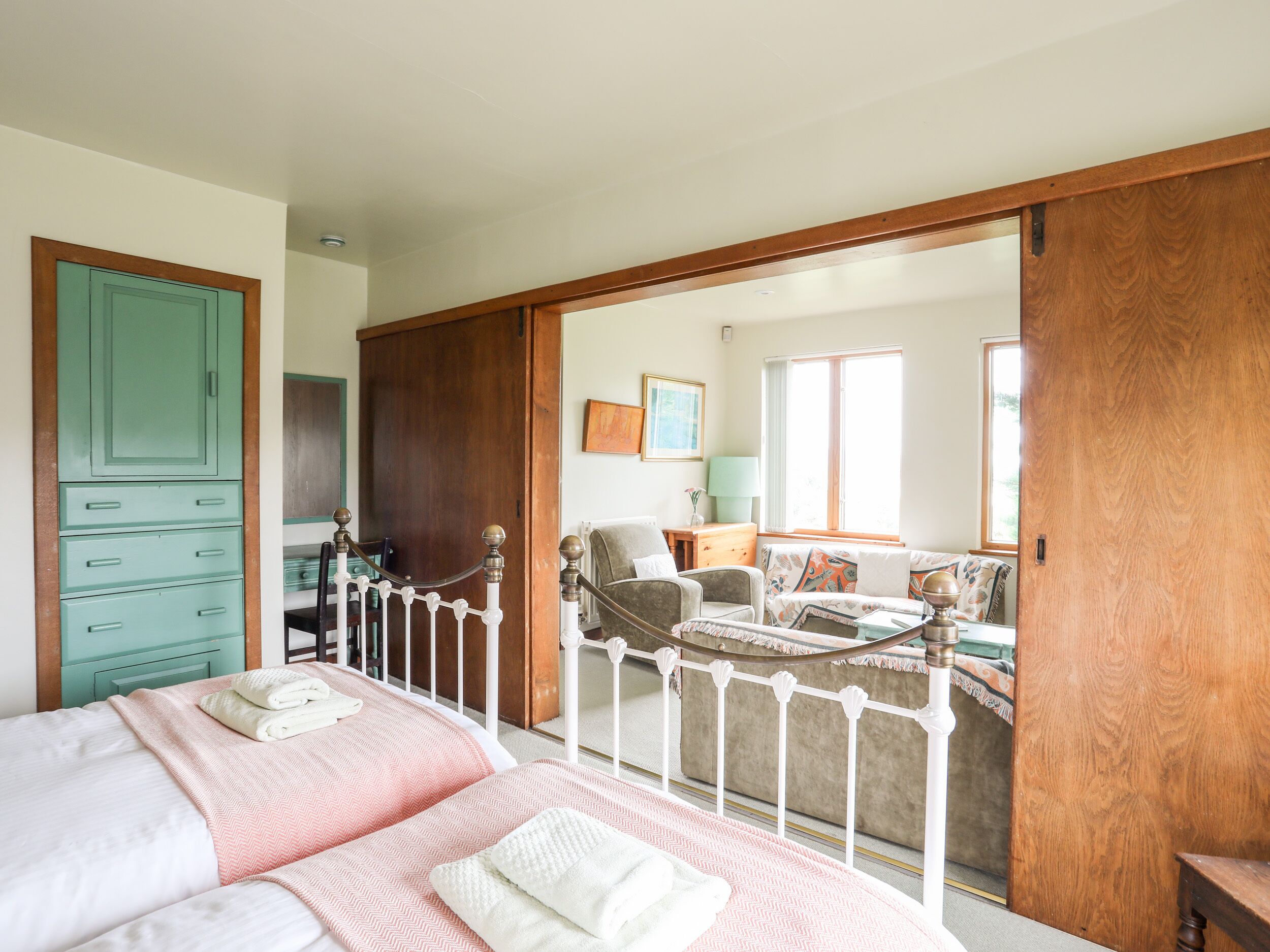 A bedroom with a bed, a wardrobe, and a view into a living room with a sofa and a table.