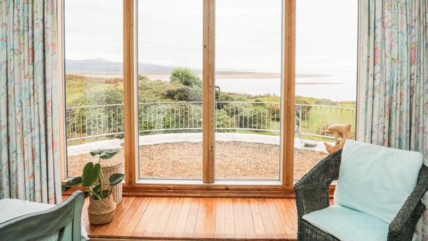 A room with a large sliding glass door leading to a balcony with a view of a lake and hills. - Bryn Y Mor, Borth Y Gest (Porthmadog)