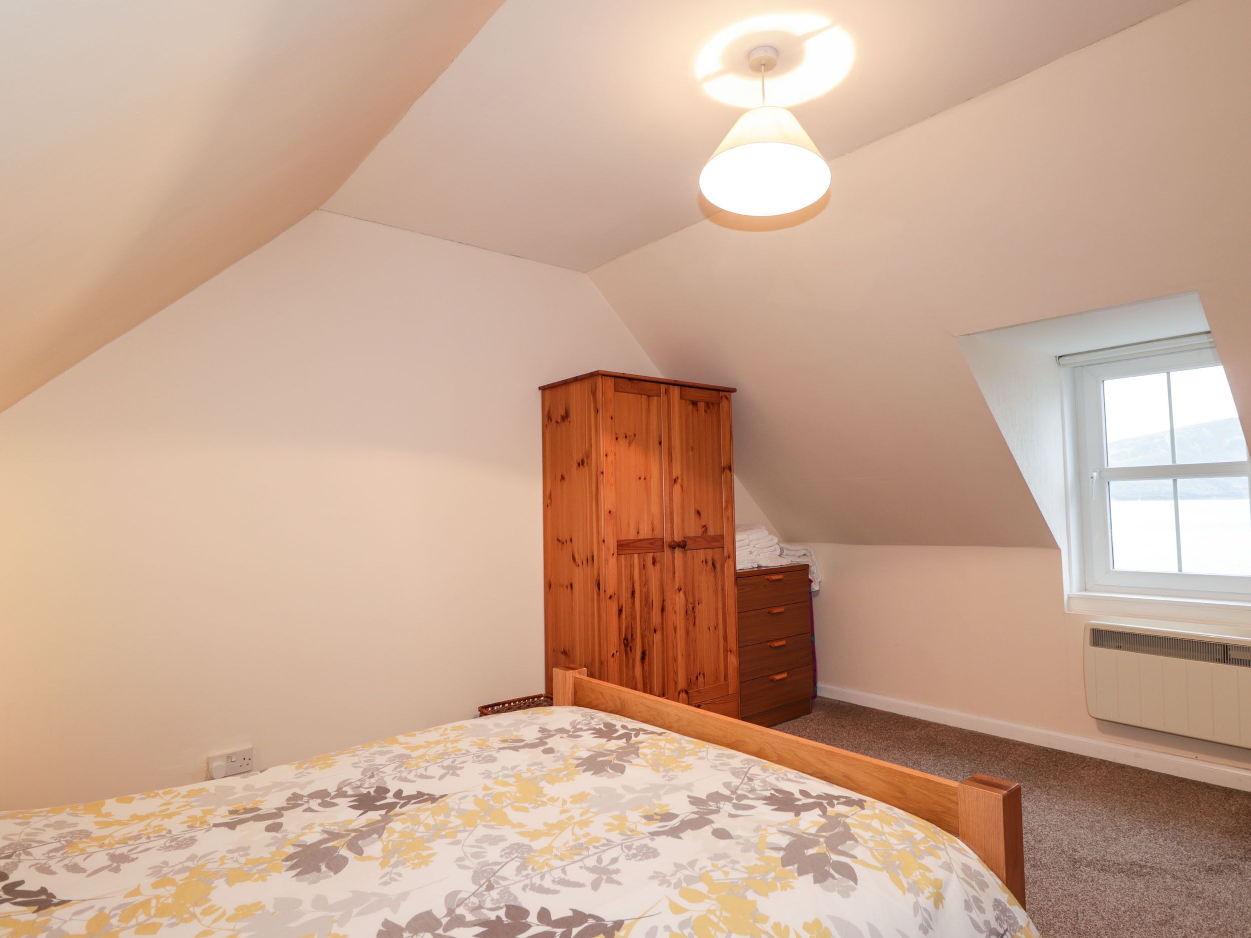 A bedroom with a sloped ceiling, a wooden wardrobe, a bed with a floral bedspread, and a window with a view.
