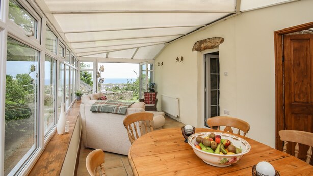 A covered patio with a wooden table and chairs, a bowl of fruit, and a view of the sea. - Fron Fawr (Pwllheli)