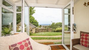 A conservatory with a view of a rural landscape, including a stone wall and a thatched-roof building. - Fron Fawr (Pwllheli)