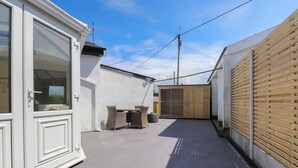 Un patio exterior estrecho con una puerta blanca, una valla de madera y suelo de baldosas.