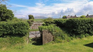 Un paisaje rural con un muro de piedra, una verja metálica y casas al fondo.