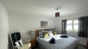 A neatly arranged bedroom with a bed, a chair, a nightstand, a lamp, and a window with curtains. - Pier View (Dunoon)