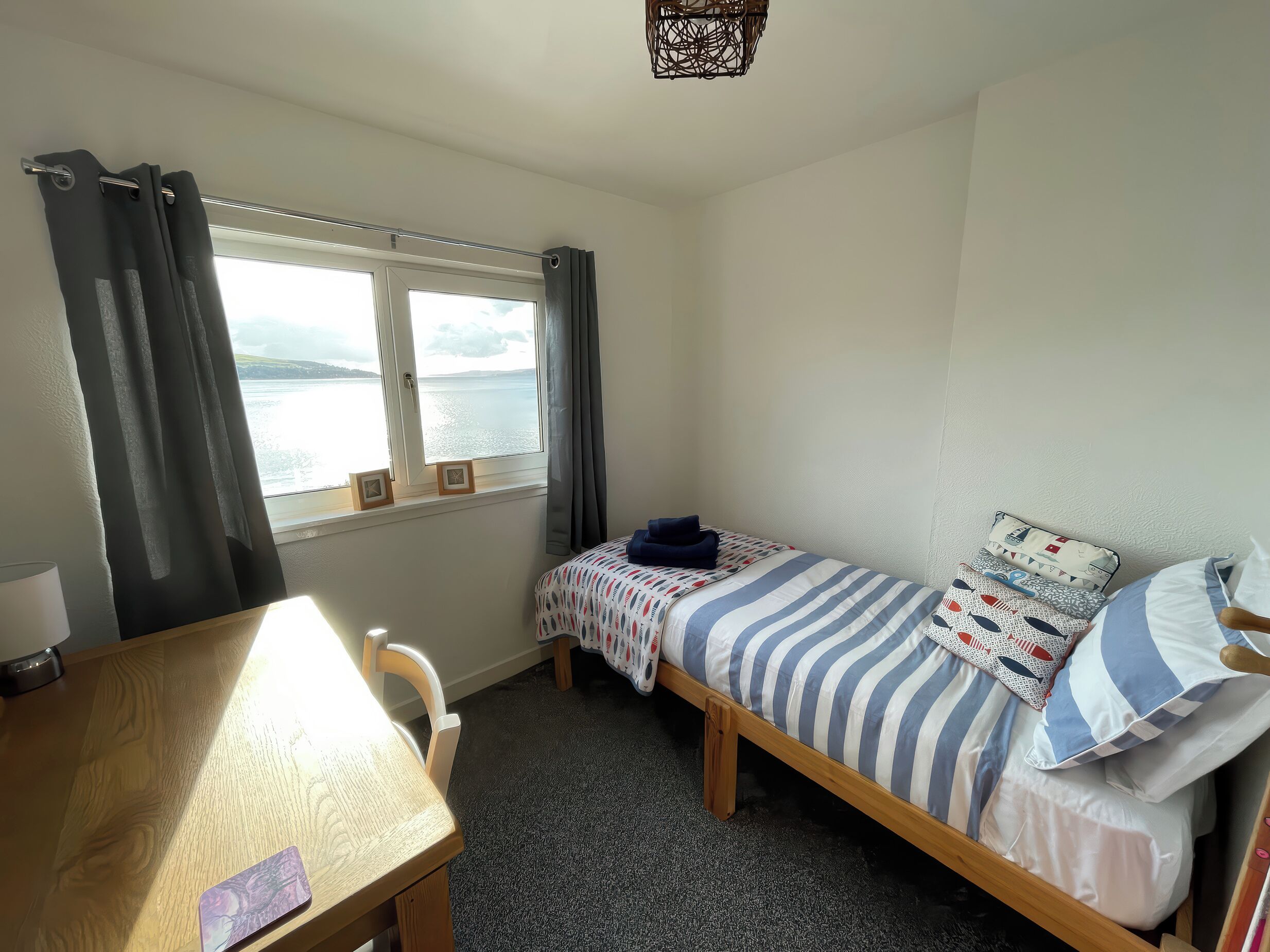 A small, tidy room with a wooden desk, a single bed with striped bedding, and a window offering a view of a body of water.
