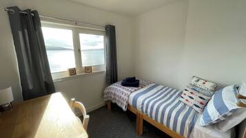 Quarto pequeno e arrumado, com escrivaninha de madeira, uma cama simples com roupa de cama listrada e uma janela com vista para um corpo d'água.