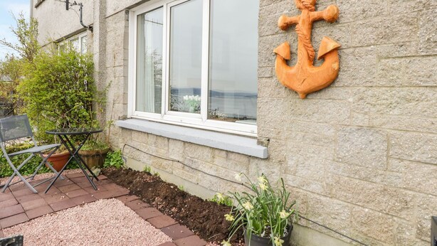 A building with a stone exterior, a large anchor sculpture on the wall, a window with white frames, and a small outdoor seating area with a table and chairs. - Pier View (Dunoon)