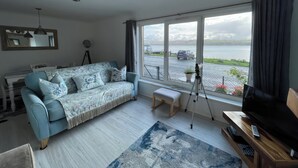 A living room with a sofa, a television, and a large window overlooking a body of water. - Pier View (Dunoon)
