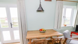 A dining area with a wooden table, two chairs, a bowl of fruit, a hanging lamp, and a window with curtains. - Wigrams Canalside Cottage (Southam)