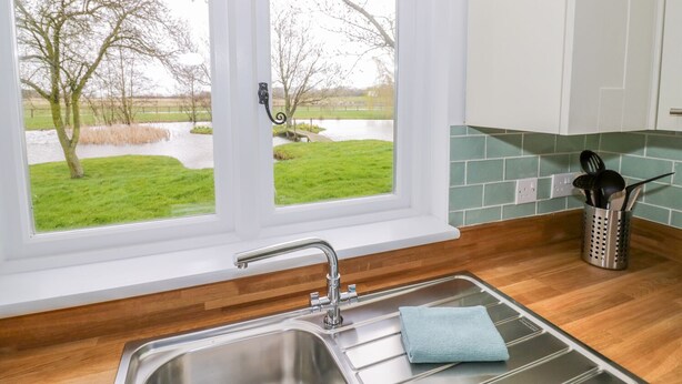 A kitchen with a double sink, a window with a view of a grassy area and trees, and a green tiled backsplash. - Wigrams Canalside Cottage (Southam)