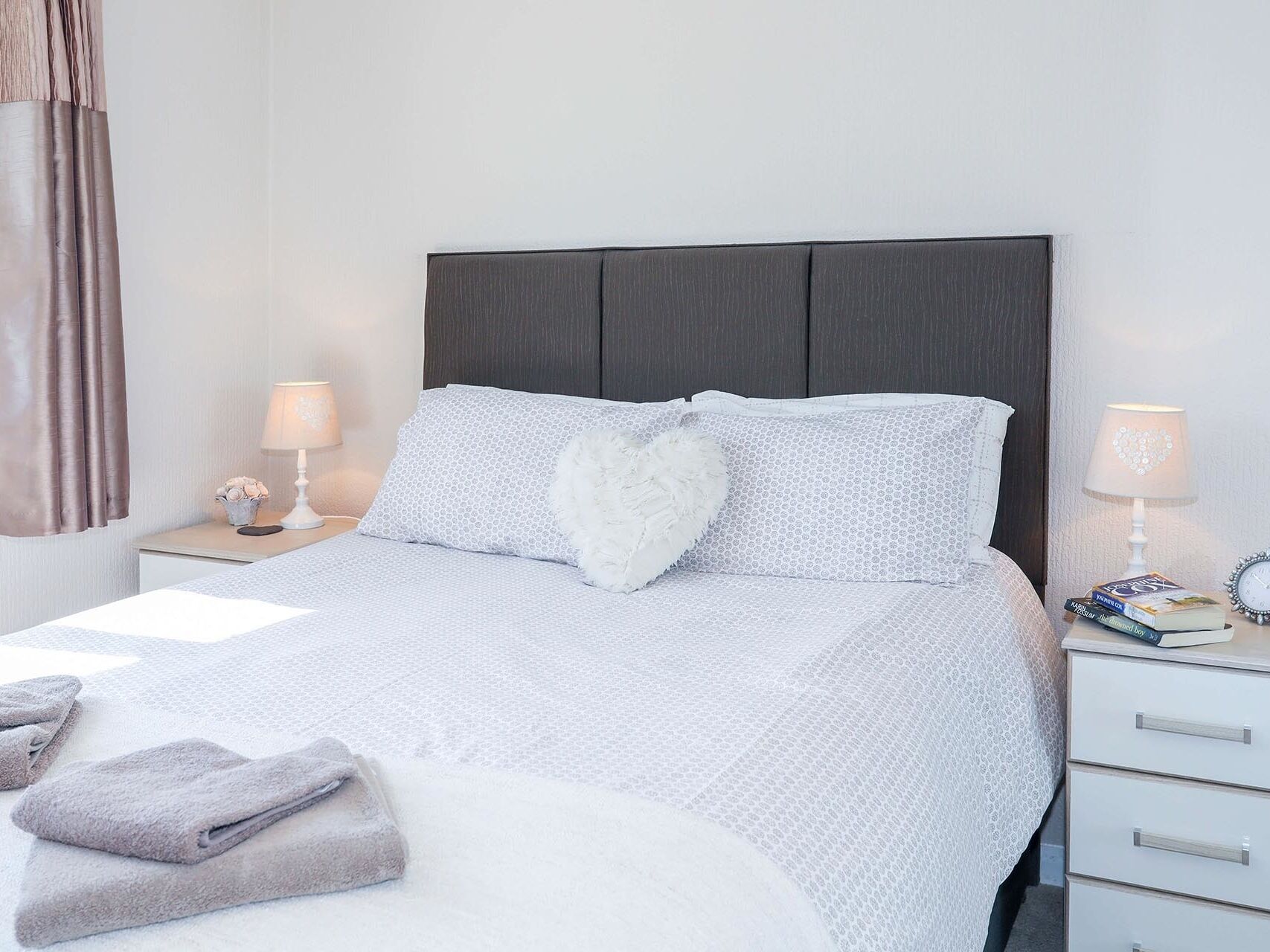 A neatly made bed with a white and grey patterned bedspread, two pillows, and a fluffy white throw. There are two bedside tables with lamps and a small clock. A stack of folded towels is visible on the bed.