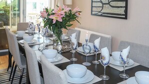 Un coin repas avec une longue table dressée pour six personnes, agrémentée d’assiettes, de bols et de verres blancs. Un vase avec des fleurs roses est posé sur la table, et un tableau orne le mur.