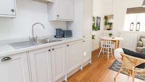 A modern kitchen with white cabinets, a stainless steel sink, and a coffee maker. There is a dining area with a round table and chairs, and a wicker chair. - 3 Poole End (Clitheroe)