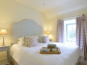 Een slaapkamer met een groot bed, een hoofdbord met bloemenprint, twee nachtlampjes, een raam met bloemen gordijnen, en een dienblad met thee en koekjes op het bed.