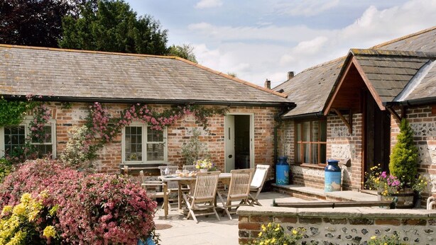 A rustic brick building with a patio area, outdoor seating, and a garden with flowering plants.