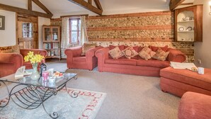 A cozy living room with a red sofa, a glass coffee table, and a patterned rug. There is a brick wall with a decorative stone pattern, wooden beams on the ceiling, and a bookshelf with books and decorative items.