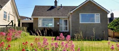 Ett tvåvåningshus med ett tak täckt av tegelpannor, en ytterdörr och ett fönster. Framför huset finns en trädgård med rosa blommor.
