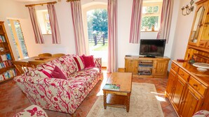 Ein gemütliches Wohnzimmer mit einer Couch, einem Couchtisch und einer TV-Konsole. Ein Bücherregal und ein Essbereich sind ebenfalls zu sehen.