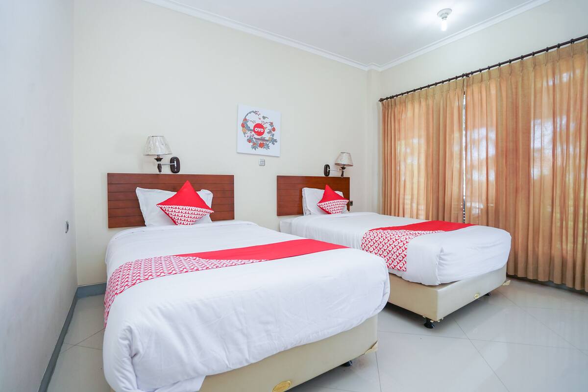 A hotel room with two beds, each with white bedding and red accents, a ceiling light, a wall clock, and a window with curtains.
