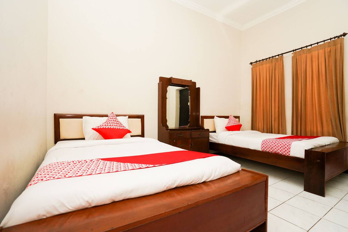 A hotel room with two wooden beds, each with a white mattress and red decorative pillows. There is a wooden nightstand with a mirror and a wooden dresser.