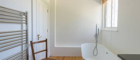 Un baño con una bañera independiente, una silla de madera, un radiador y una ventana con persianas.