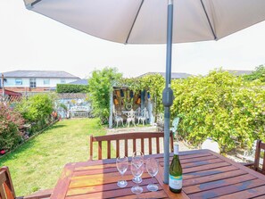 A wooden table with a bottle of wine and three glasses, under a white umbrella, in a garden with a fence and greenery.