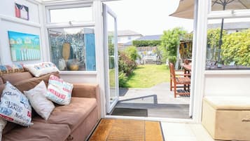 Un salon chaleureux avec un canapé marron, des coussins, une porte vitrée donnant sur un jardin et un coin repas avec une table et des chaises.