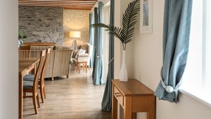 A cozy living room with a stone wall, wooden ceiling beams, a dining area with wooden chairs, a small table, and a potted plant. - Tyn Towyn - Ty Coch (Holyhead)
