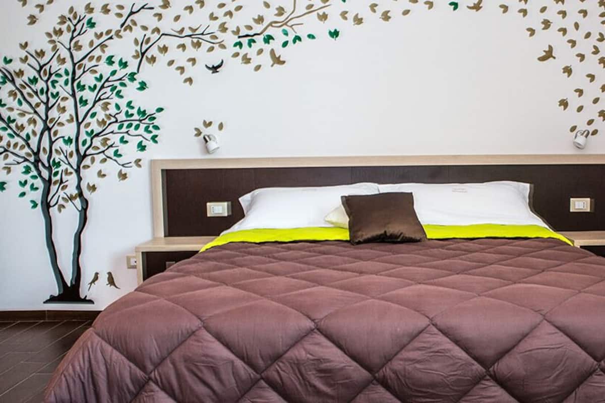 A neatly made bed with a brown quilted bedspread, a white pillow, and a brown accent pillow. There is a wall-mounted shelf with a vase and a small plant.