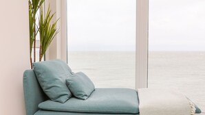 A teal sofa with two cushions and a throw blanket, positioned next to a window overlooking the sea. - Seasound (Ilfracombe)
