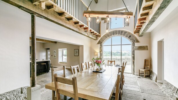 A spacious dining area with a long wooden table and chairs, a stone floor, and a view through an arched window. - Top Barn (Sowerby Bridge)