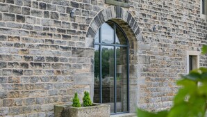 A stone building with a large arched window and a small planter with two shrubs. - Top Barn (Sowerby Bridge)