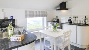 A modern kitchen with a white table, chairs, a window with a view, a black kettle, and a bottle of Hardyi on the counter. - Amolca (Amlwch)