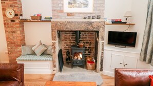 A cozy living room with a brick fireplace, a television mounted on the wall, and a comfortable seating area with a sofa and a bench. - Wyebrow Cottage (Bakewell)
