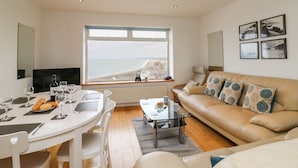 A modern living room with a large window offering a view of the sea and cliffs, a dining area with a round table and chairs, a sofa with patterned cushions, and a coffee table. - The View (Portland)