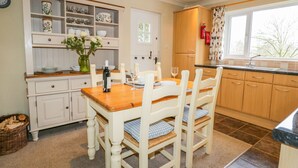 A cozy kitchen with a wooden table and chairs, a built-in shelf with dishes, and a window with a view. - Kirkstone Cottage (Ambleside)