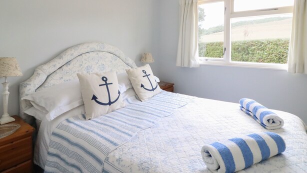A neatly made bed with striped bedding, two pillows with anchor designs, a window with curtains, and a view of greenery outside. - Woodlands Cottage (Dorchester)