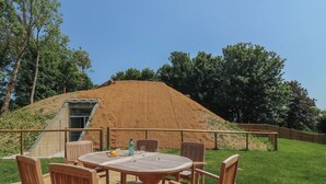 An outdoor patio with a round wooden table and chairs, a small building with a sloped roof, and a grassy area with trees in the background. - Woodlands Cottage (Dorchester)