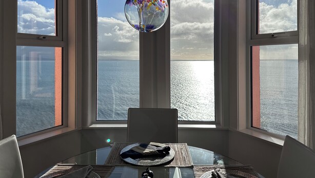 Una habitación con un ventanal grande con vista al mar, una mesa de vidrio con posavasos y una silla.